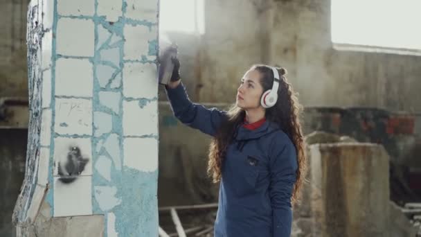 魅力的な女の子は、エアゾール塗料を使用して断念された建物に落書きに古い汚れた列を飾ることに集中されます。若い女性は、ヘッドフォンで音楽を聴く. — ストック動画