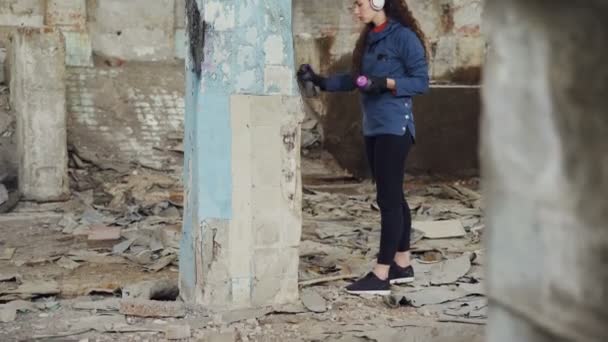 Κλίση-up shot slim κορίτσι σύγχρονη ζωγράφου κατάρτιση γκράφιτι σε υψηλή στήλη σε ερειπωμένη αποθήκη με σπρέι βαφής και να ακούτε μουσική με ακουστικά. — Αρχείο Βίντεο