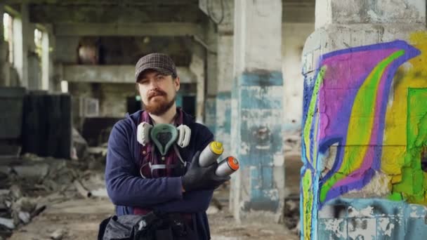 Портрет бородатого хлопця графіті, що стоїть у старій порожній будівлі з графітним живописом на задньому плані і тримає розпилювальну фарбу. Чоловік має газову маску і носить рукавички . — стокове відео