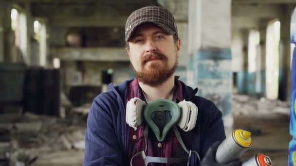 Retrato de primer plano de un guapo artista de graffiti barbudo parado dentro de un edificio abandonado usando gorra, guantes y respirador y sosteniendo pintura en aerosol — Vídeo de stock