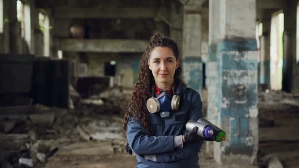 Retrato de sorrindo bela garota pintor de grafite em pé no antigo edifício abandonado, segurando tintas spray e olhando para a câmera. Mulher tem máscara protetora e luvas . — Vídeo de Stock