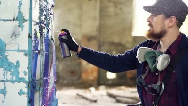 Serious man is painting graffiti on dirty pillar in old empty house with aerosol paint. Guy is wearing casual clothes and trendy cap, he has protective gloves and mask. — Stock Video