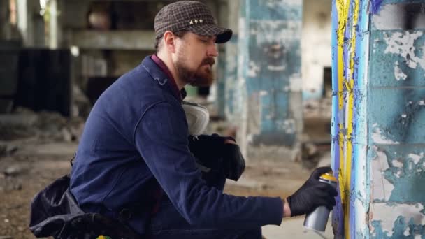 Hombre adulto serio artista de graffiti en guantes de cuero está pintando en pilar dentro dañado edificio industrial vacío y en cuclillas. Concepto de creatividad y personas . — Vídeo de stock