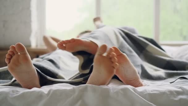 Husband and wife are lying in bed under blanket and talking then hugging, focus on bare feet touching each other. Romantic relationship and loving people concept. — Stock Video