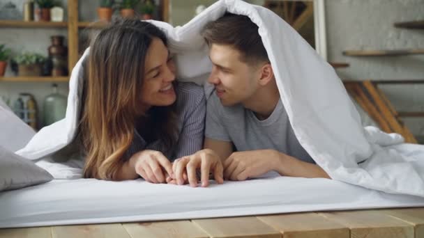 Liefdevol paar vriendin en vriend en liggend in bed onder deken praten, lachen en kussen tijdens gelukkig weekend thuis. Relatie en mensen concept, — Stockvideo