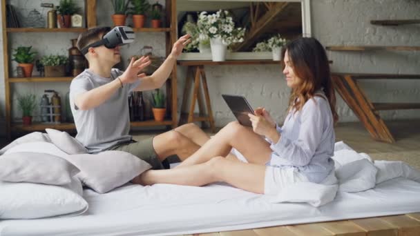 Tânărul folosește ochelari de realitate virtuală în timp ce soția lui atrăgătoare atinge ecranul tabletei, se uită la soțul ei și zâmbește. Tehnologie modernă și concept de relație . — Videoclip de stoc