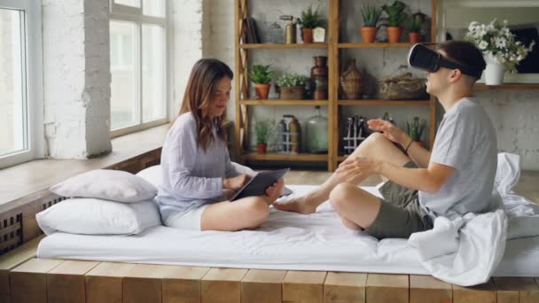 Young couple is having fun man is wearing virtual reality glasses while woman is touching tablet screen, they are talking and laughing sitting on double bed in bedroom. — Stock Video