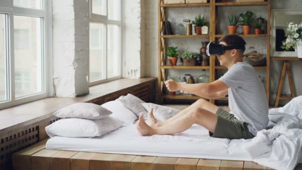 Jovem feliz em óculos de realidade virtual está sentado na cama e mãos em movimento como se ele está dirigindo carro volante girando, cara está desfrutando de experiência e rindo . — Vídeo de Stock