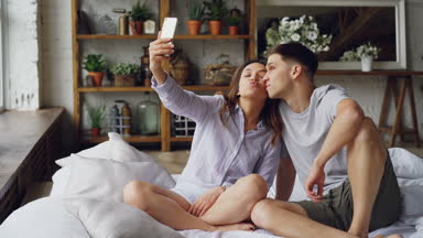 Sevgi dolu çift selfie kameraya bakarak, poz ve birlikte yatağa evde otururken komik surat smartphone ile alıyor. Teknoloji ve ilişki kavramı.