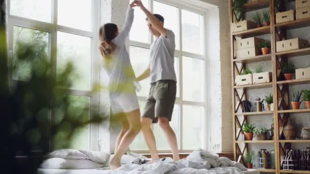 Jong getrouwd paar is springen en dansen op tweepersoonsbed in lichte kamer met grote ramen, gelukkige mensen zijn met plezier en lachen. Liefde, entertainment en mensen concept. — Stockvideo