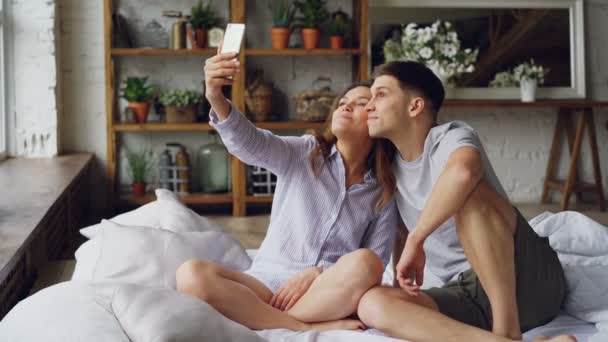 Alegre pareja amorosa está tomando selfie con teléfono inteligente mirando a la cámara, posando y haciendo caras divertidas mientras se sientan juntos en la cama en casa. Concepto de tecnología y relación . — Vídeo de stock