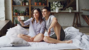 Sevimli Genç çift smartphone ile görüntülü görüşme yapıyor, gençler konuşuyor ve çift kişilik yatak yatak odasında otururken el hareketi. Teknoloji ve iletişim kavramı.