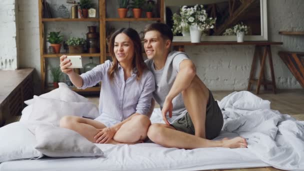 Очаровательная молодая пара делает видеозвонок со смартфона, молодые люди разговаривают и жестикулируют, сидя на двуспальной кровати в спальне. Технология и коммуникация . — стоковое видео