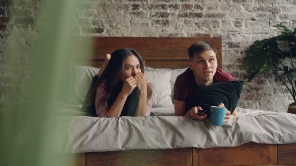Mladý muž se dívá na televizi se svou přítelkyní, ležící na posteli a drží vzdálenou, drží polštáře a hrnek. Televize, rodinný život a zábavu koncepce. — Stock video