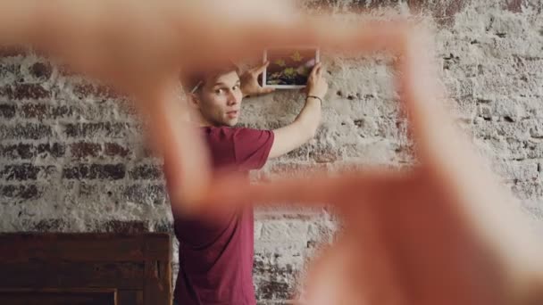 Cheerful man is choosing place for modern picture on brick wall while his wife is making frame shape with her fingers and looking at him, female hands in foreground. — Stock Video