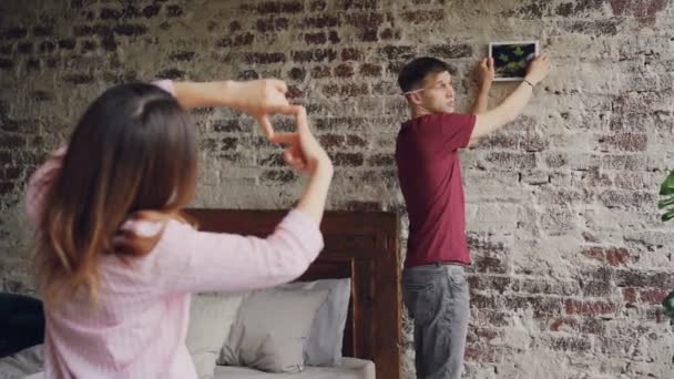 Przystojny facet jest zbieranie się na mur z cegły powiesić nowoczesny obrazek, podczas gdy jego dziewczyna jest Dokonywanie ramki z jej palce i gestykulacji, zatwierdzające jego decyzji z kciuk. — Wideo stockowe