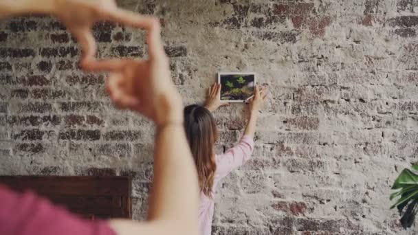 Mujer asiática bonita está eligiendo el lugar para la fotografía enmarcada en la pared de ladrillo, mientras que su novio está haciendo la forma del marco con los dedos y mirándola, las manos en primer plano . — Vídeos de Stock