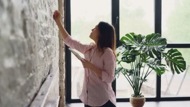 Mujer creativa está decorando su apartamento estilo loft elegir lugar en la pared de ladrillo para la imagen hermosa y el lugar de marcado con lápiz. Creatividad y concepto interior . — Vídeo de stock