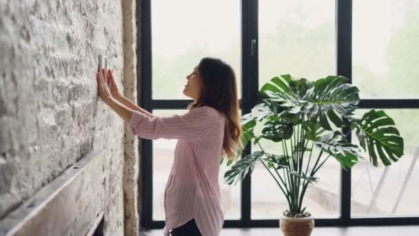 Bonita morena propietario de la casa está decorando su habitación estilo loft colgando imagen en la pared de ladrillo lugar de elección. Diseño de interiores, tareas domésticas, decoración de apartamentos y concepto de personas . — Vídeos de Stock