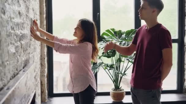 Cheerful young woman is choosing place for framed photograph on brick wall while her husband is helping her, happy couple is talking and making decision. — Stock Video