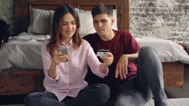 Молодая женщина держит банковскую карточку и делает онлайн-платеж со смартфона, а затем целует своего парня, сидя вместе на полу спальни. Концепция банковского дела и людей . — стоковое видео