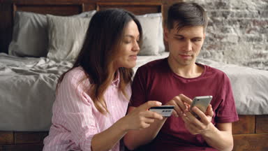 Yakışıklı genç adam onun çekici karısı onun banka kartı tutan ve onunla konuşmaya yanında oturuyor smartphone kullanarak Internet alımları için ödüyor.