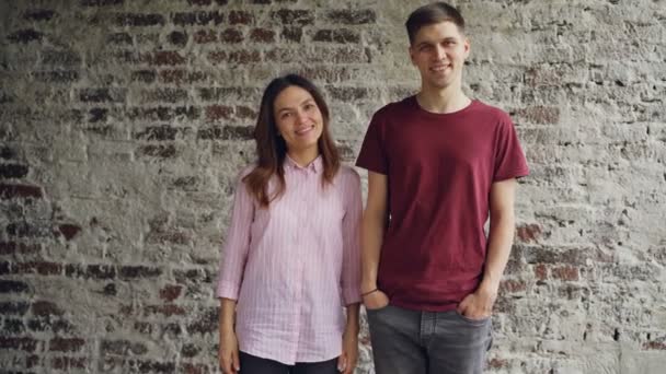 Portrait of attractive mixed race couple standing together against brick wall looking at camera and smiling. Happy people and modern interior concept. — Stock Video