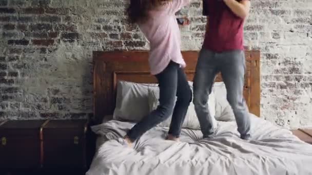 Verspielte junge Leute tanzen auf dem Bett, hören Musik und singen mit Föhn und Fernbedienung als Mikrofone, die zusammen Spaß haben. — Stockvideo