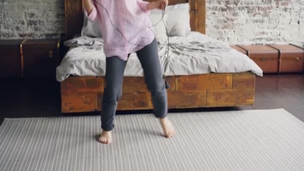 有魅力的年轻女子在衬衫和牛仔裤是跳舞和唱歌举行吹风机和使用它作为麦克风在尼斯阁楼式卧室附近的现代双人床. — 图库视频影像