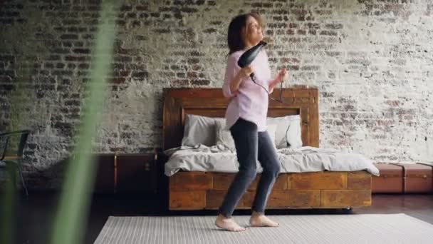 Chica divertida está bailando y cantando sosteniendo secador de pelo divertirse en el dormitorio en la alfombra cerca de la cama doble. Mujer joven activa está disfrutando de tiempo libre y escuchar música . — Vídeos de Stock
