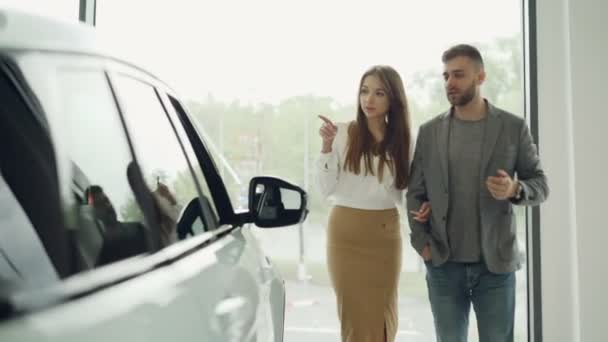 Atractivo joven pareja confiado barbudo chico y su novia bonita están eligiendo nuevo automóvil juntos mirando coche de lujo en la concesionaria de automóviles y hablando . — Vídeo de stock