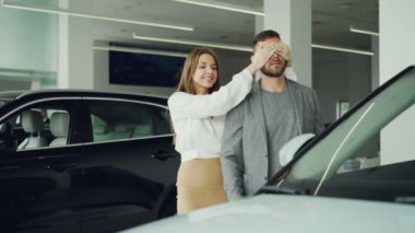 Genç ve güzel kadın koca gözleri kapanıyor ve onu yeni araba otomobil Showroom için lider, heyecanlı adam gülüyor, Öpüşme ve eşi sarılma.