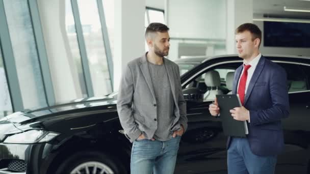 El chico guapo está hablando con el gerente de concesionarios de automóviles en el espectáculo de automóviles discutiendo el nuevo modelo de automóvil, distribuidor está sosteniendo documentos y pluma. Concepto de compra de vehículos y personas . — Vídeos de Stock