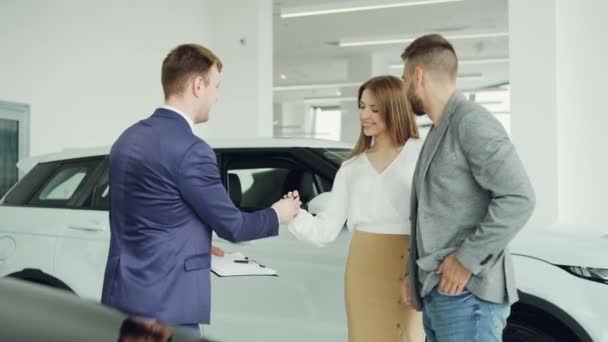 Fröhliche Autohaus Verkäufer schüttelt Hände mit jungen Käufer schöner Mann gibt dann Schlüsselanhänger an seine glückliche Frau, Paar umarmt und küsst. — Stockvideo