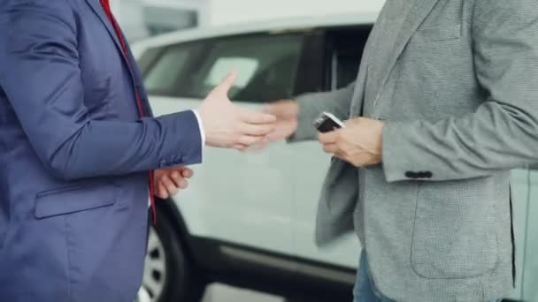 Male worker of car showroom is giving car keys to buyer young man and shking hands with him standing beside luxurious new car. Selling and buying vehicles concept. — Stock Video