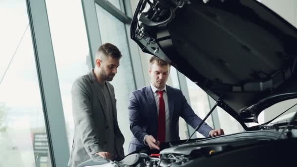 Ervaren autohandelaar is nieuwe klant motor onder geopende motorkap tonen en hem te vertellen over de kwaliteit en de kenmerken van auto model. Controle auto kwaliteit concept. — Stockvideo