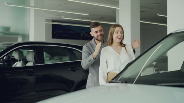 Neşeli adam karısını elleriyle gözlerini kapatmak için sürpriz yapıyor ve onu lider için güzel yeni araba motor showroom, mutlu kadın sarılma ve onu öpmek. — Stok video