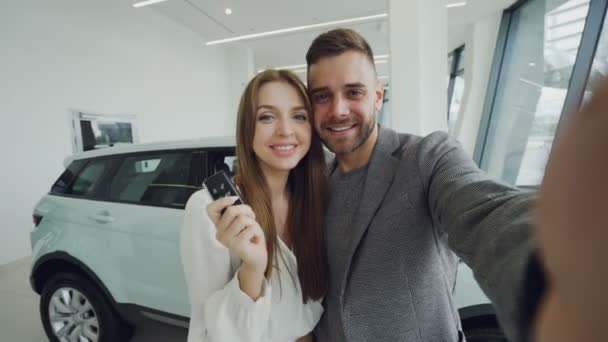 Atractiva pareja joven se está tomando selfie con las llaves del coche en la sala de exposición del motor moderno después de comprar un automóvil nuevo. Están mirando a la cámara, besando y sonriendo . — Vídeos de Stock
