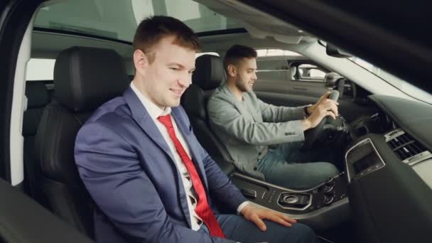 Comprador de carro cara alegre está falando com vendedor profissional sentado dentro caro auto e tocando painel de instrumentos e volante. Conversação e conceito de test drive . — Vídeo de Stock