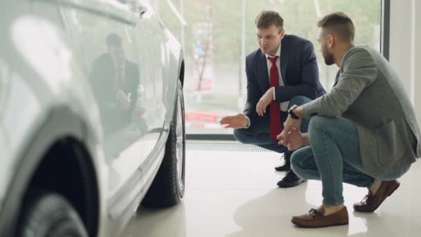 Dos hombres cliente alegre y vendedor amistoso están hablando y haciendo gestos sobre el modelo de coche mientras se agacha cerca de automóviles caros. Concepto de compra y venta de vehículos . — Vídeos de Stock