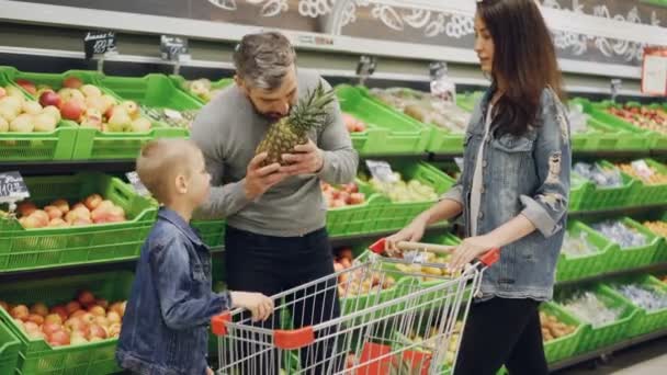 Отец, мать и ребенок выбирают ананас в продуктовом магазине, трогают и нюхают его, показывая большой палец вверх. Видны полки с вкусными фруктами и тележка для покупок . — стоковое видео