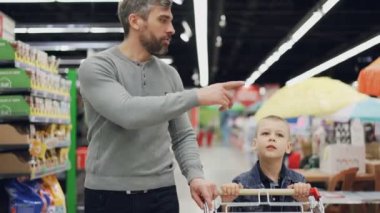 Neşeli adam neşeli onun çocuğuna alışveriş, arabası bakarak ürünler işaret, söz ve gülüyor, süpermarkette alışveriş ile yürüyorlar.