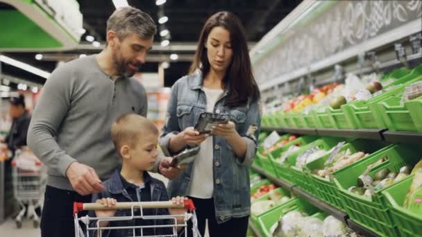 Bonito barbudo cara, sua esposa atraente e filho bonito estão escolhendo legumes em bandejas no supermercado, falando, rindo e colocando produtos no carrinho . — Vídeo de Stock