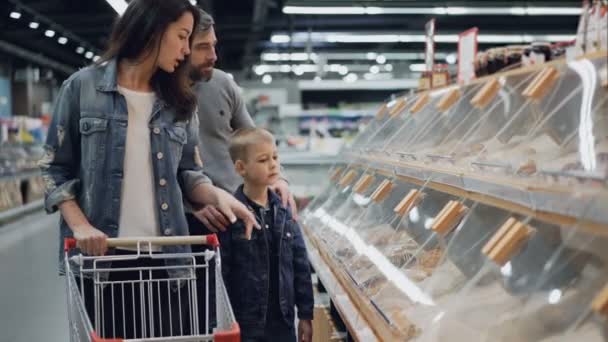 Mère, père et leur fils achètent de la nourriture dans un supermarché en pointant du doigt des boîtes en plastique sur des étagères et en parlant. Grand magasin d'alimentation et ses employés sont visibles en arrière-plan . — Video