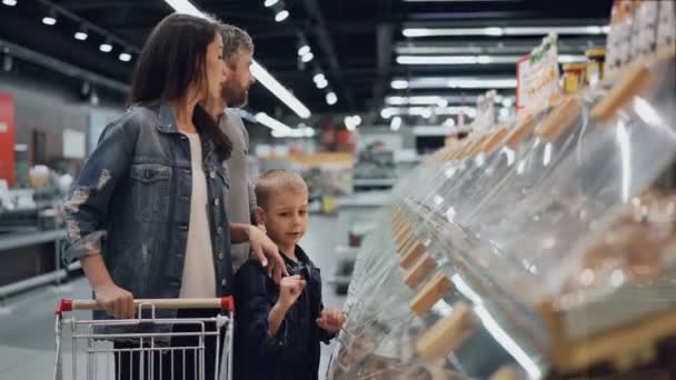 Gelukkige familie moeder, vader en schattige jongen zijn kiezen van voeding in grote supermarkt, wijzend op plastic verpakkingen, praten, lachen en lachen. Mensen en shopping concept. — Stockvideo