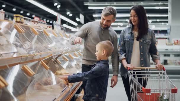 La jeune famille choisit le pain dans le département de boulangerie au supermarché, le petit garçon prend le pain du récipient en plastique et le sent puis le met dans le chariot . — Video