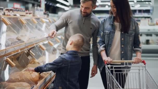 Il figlio utile sta aiutando i suoi genitori a comprare il pane, il padre sta aprendo il contenitore e il ragazzo sta prendendo il pane e lo annusa e poi lo mette nel carrello. Shopping insieme concetto . — Video Stock