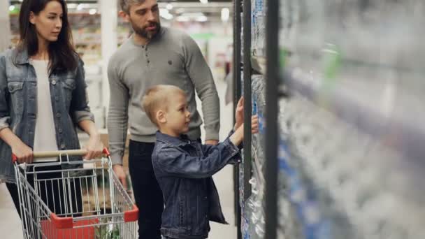 かわいい子はスーパー マーケットの棚からの水のボトルを撮影と買い物カゴに入れて、彼の愛情深い両親見ている彼は、笑顔でお返事. — ストック動画