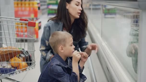 Молодая привлекательная женщина и ее милый блондин сын выбирают еду в супермаркете указывая на продукты и говорить. Видна тележка с яркими продуктами . — стоковое видео