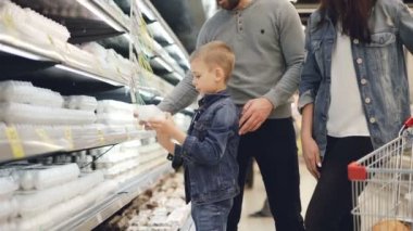 Sevimli küçük çocuk hipermarket, bakarak yumurta tepsi raf alıyor ve yumurta sayım, gülümseyen ailesi karton arabası alışveriş ve konuşmaya koyarak.